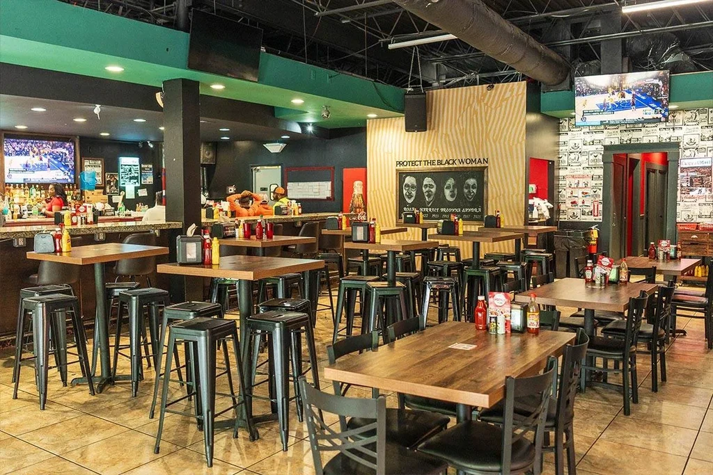 Casual industrial bar interior; "PROTECT THE BLACK WOMAN" sign, sports TVs, patrons.