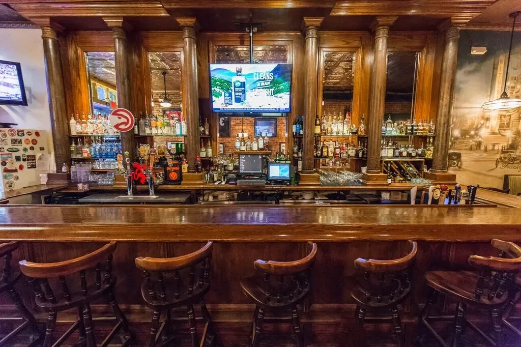 Classic wooden bar counter with liquor bottles, multiple TVs, and stools.