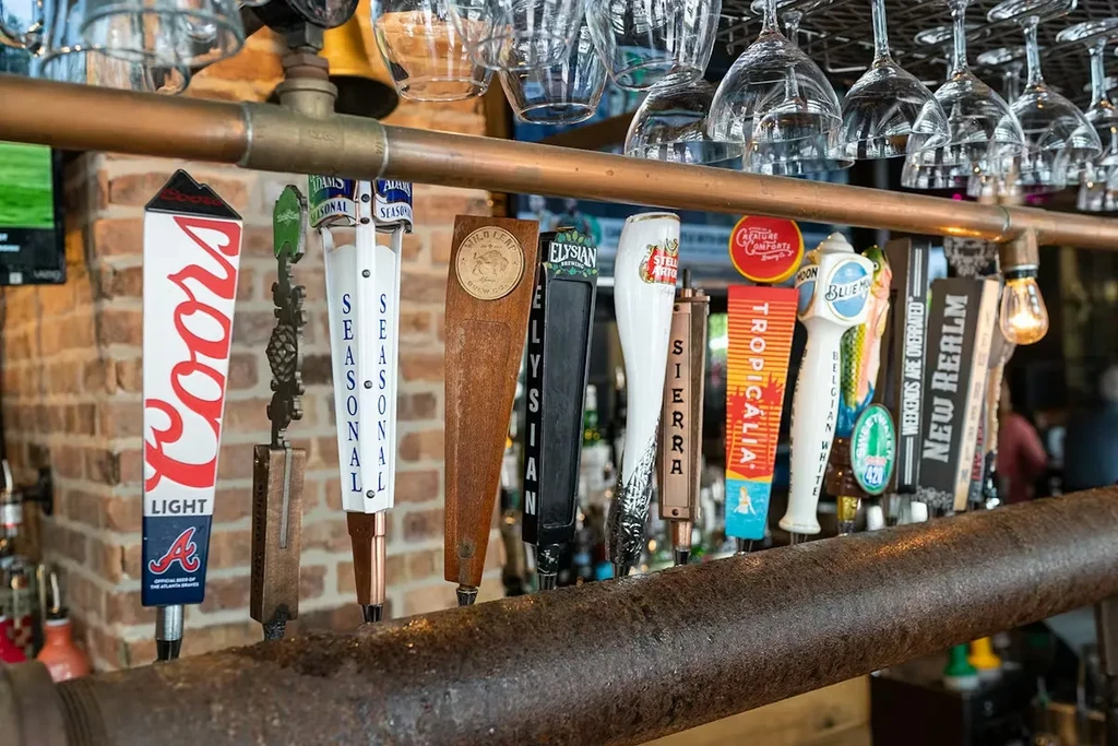 Close-up of diverse beer tap handles, hanging glasses, and a copper pipe.