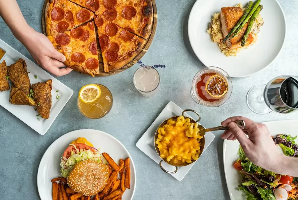 Overhead table with pizza, salmon, burgers, drinks; hands reaching for food.