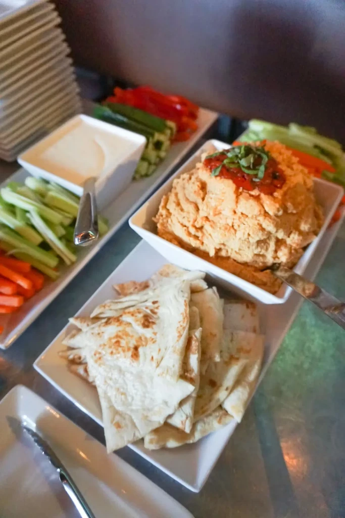 A buffet with hummus, pita triangles, vegetable sticks, and two dips.