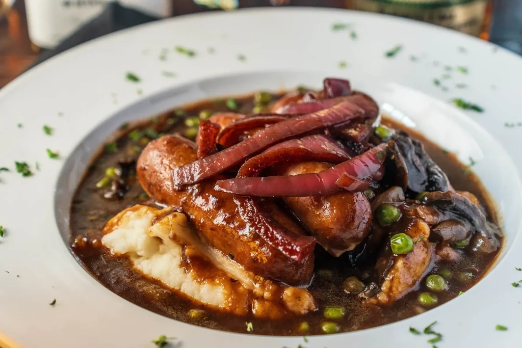 Plate of sausages, mashed potatoes, gravy, braised onions, peas, and herbs.