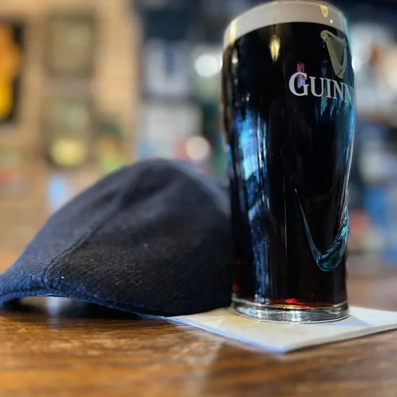 Guinness pint and dark flat cap on a warm wooden table.