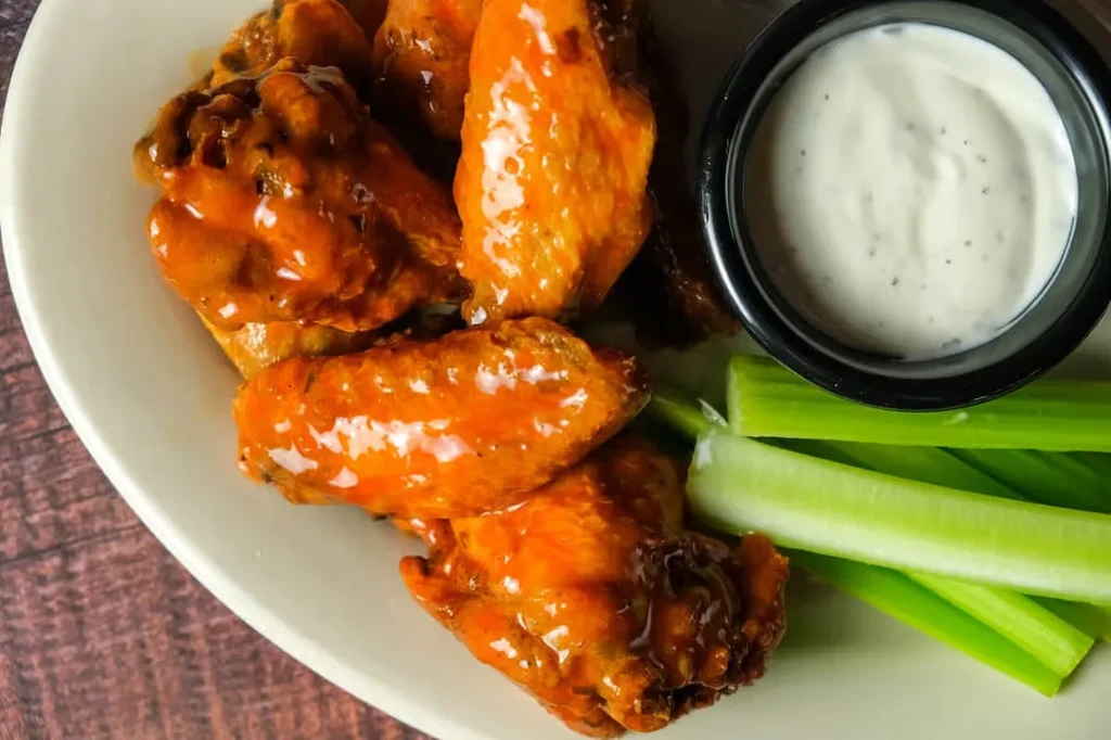 Plate of glossy orange buffalo wings, white dipping sauce, and celery sticks.