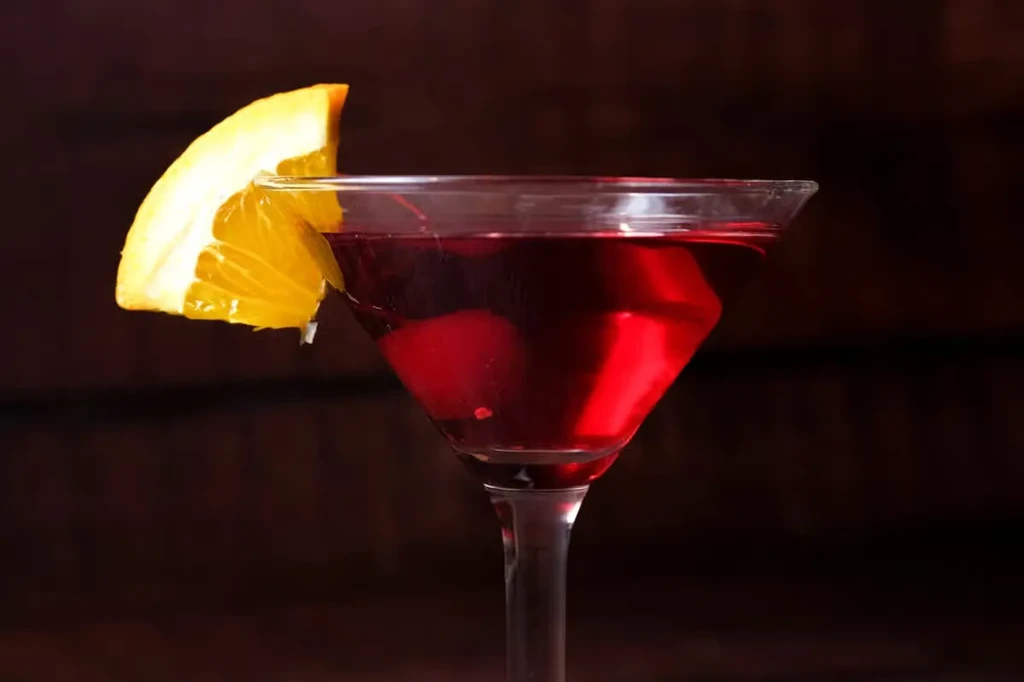 Vibrant red cocktail in martini glass, garnished with orange wedge.