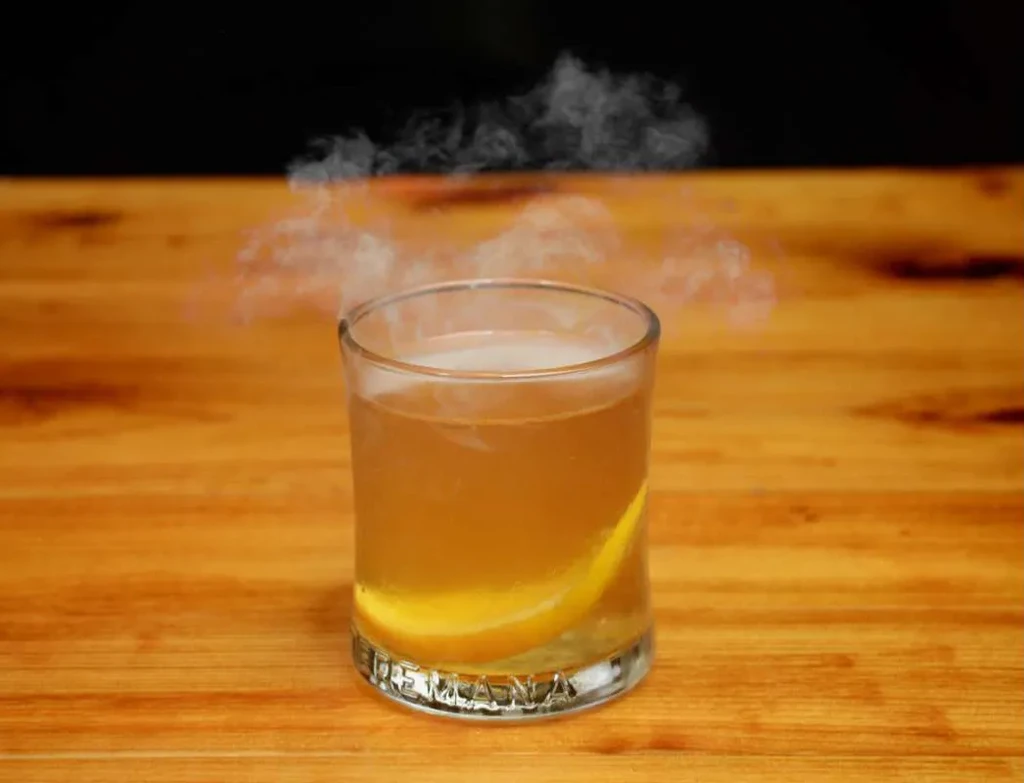 Clear glass of steaming amber liquid with citrus on wood.