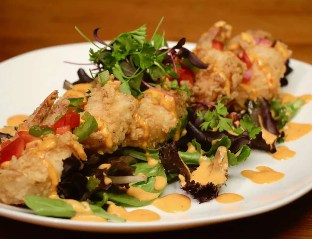 Fried shrimp, orange sauce, mixed greens, peppers, herbs on white plate.