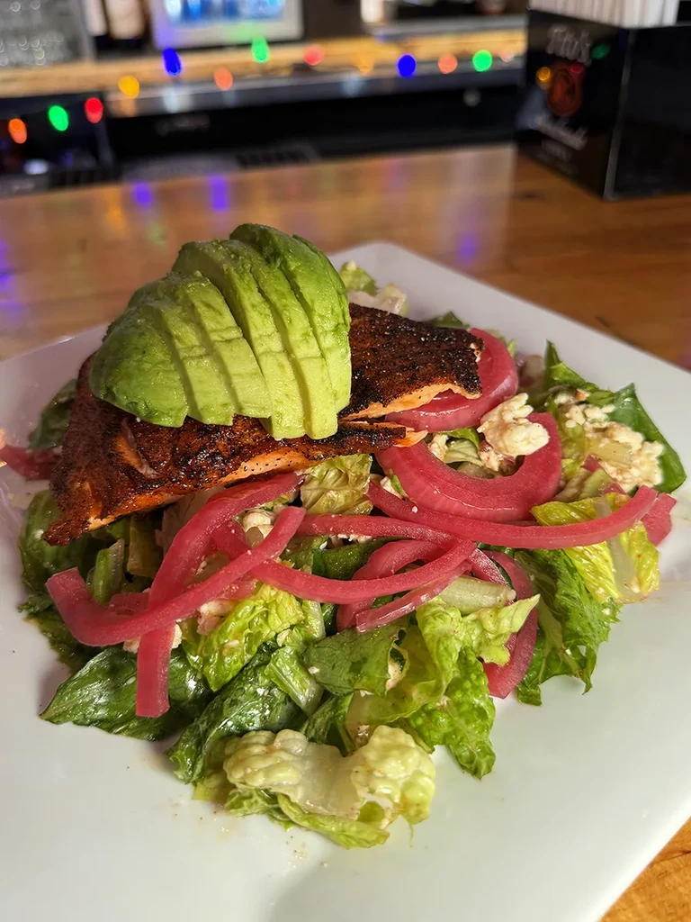 Close-up of a plated salad with seared fish, avocado, lettuce, feta, and pickled onions.
