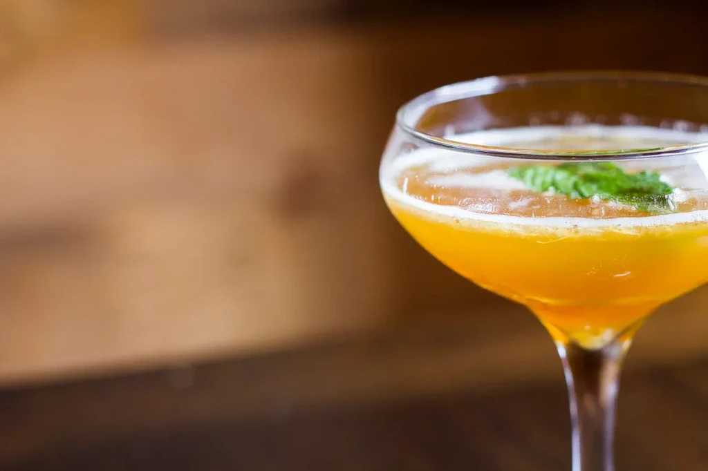 Orange cocktail in a coupe glass with foam and mint garnish.