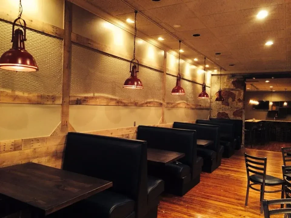 Restaurant interior with empty dark booths, rustic wood-glass wall, red lamps.