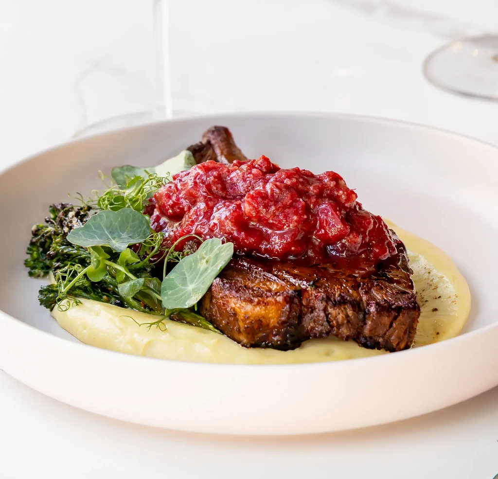 Plated meat with red sauce, broccolini, microgreens, and puree.
