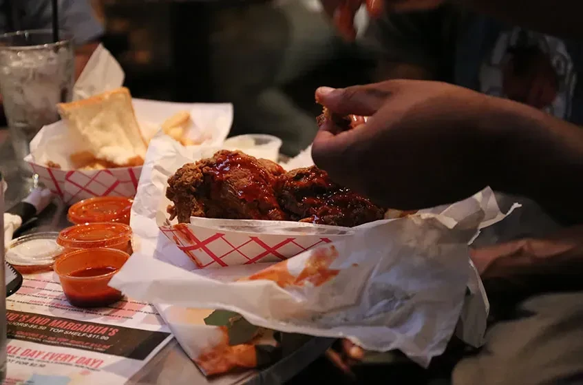 Hands holding saucy fried chicken in a checkered basket at a restaurant.