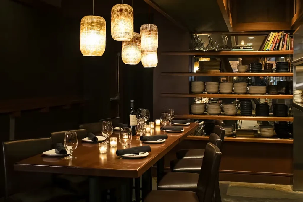 Cozy restaurant interior: long set wooden table, pendant lights, shelved dishes.