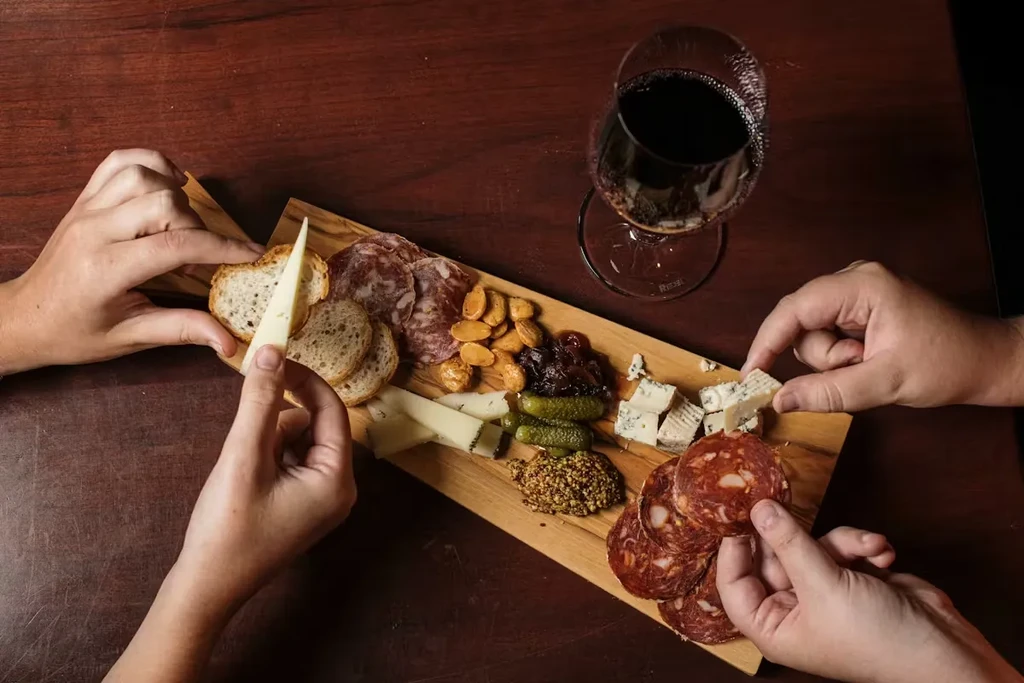 A top-down charcuterie board with meats, cheeses, and three active hands.