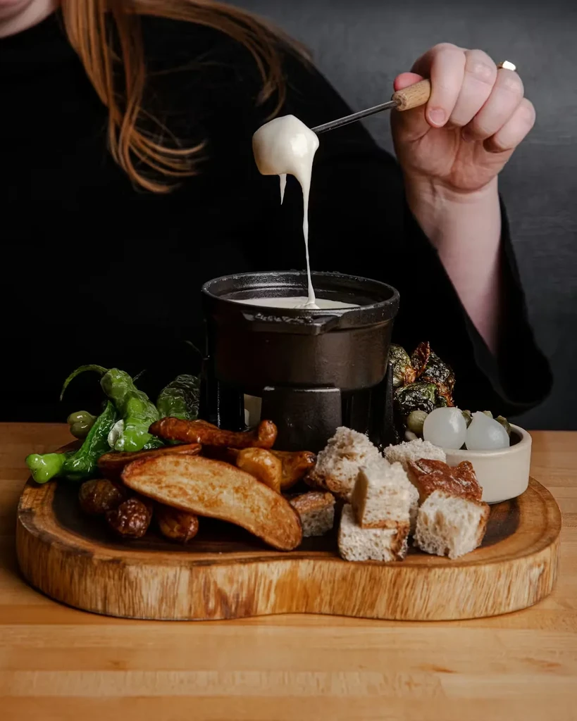 Hand dips cheese-covered food from a fondue pot with dippers.