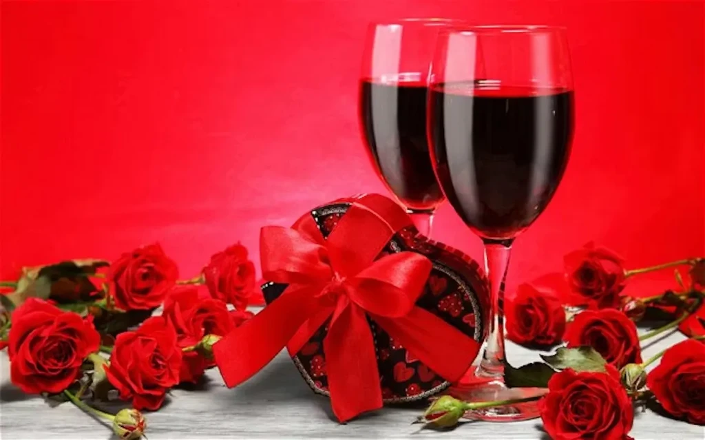 Two red wine glasses, heart gift box, and scattered roses on red.
