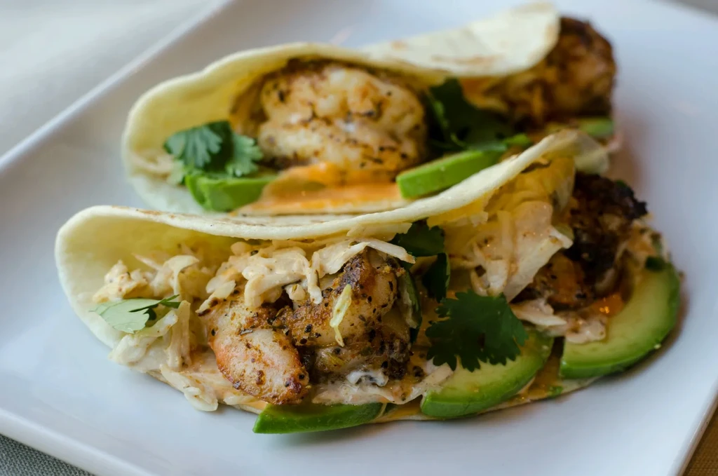 Two shrimp tacos with avocado, slaw, sauce, and cilantro on a plate.