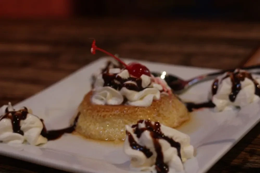 Close-up of golden flan with whipped cream, cherry, and syrup.