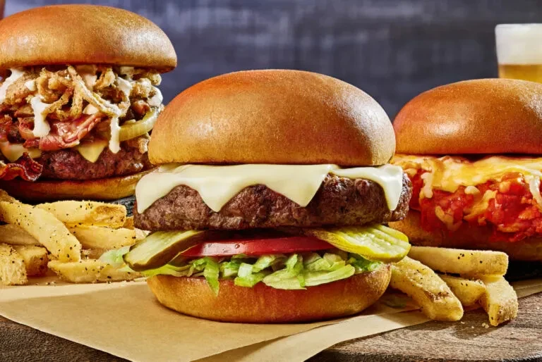 Close-up of three gourmet burgers, fries, and beer on a wooden board.