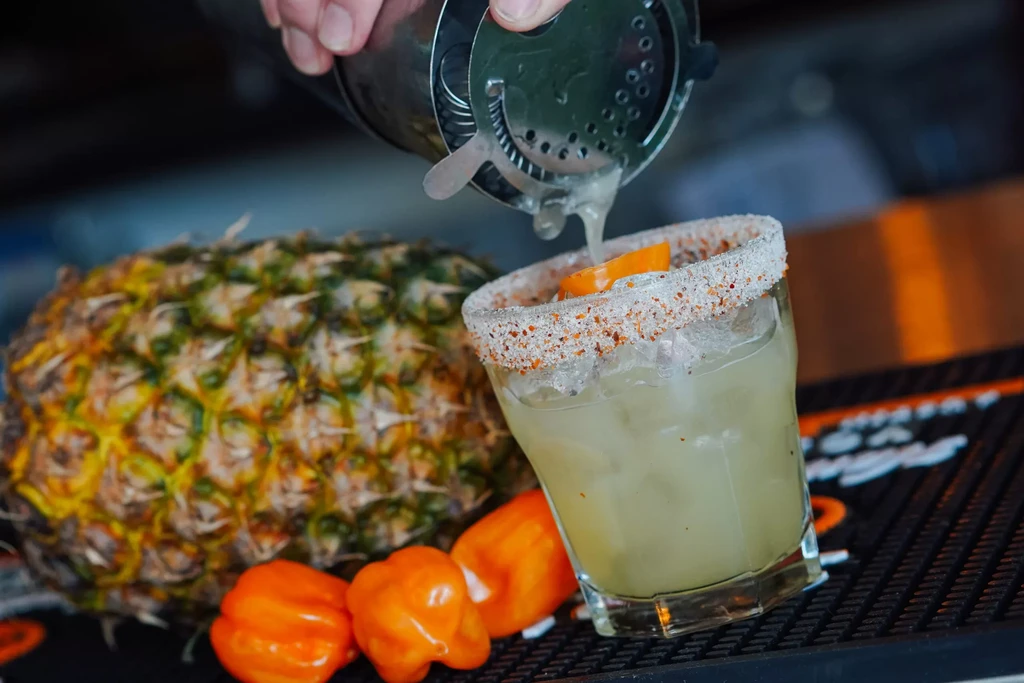 Hand pours cocktail into glass with spicy rim and peppers.