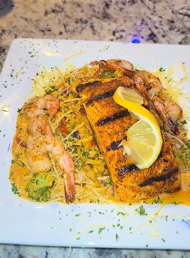 Plated grilled salmon, shrimp pasta with creamy sauce, broccoli, lemon.