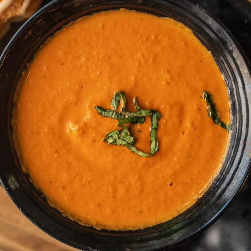 Creamy reddish-orange soup with green basil in a dark bowl.