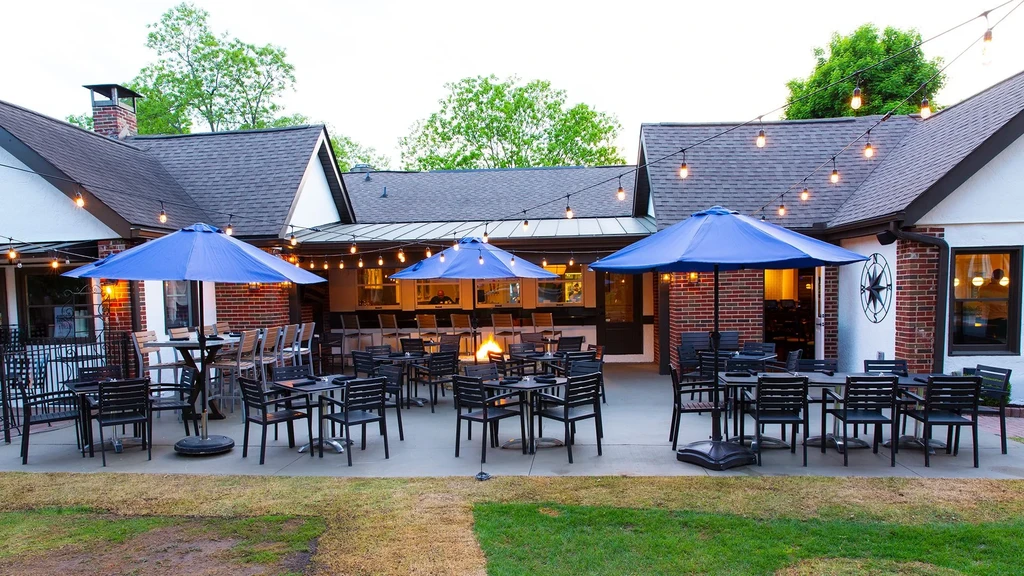 Restaurant patio with dark seating, blue umbrellas, fire pit, and string lights.