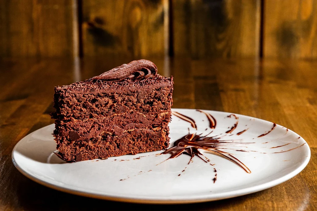 Multi-layered chocolate cake slice with sprinkles, sauce, on white plate, wooden table.