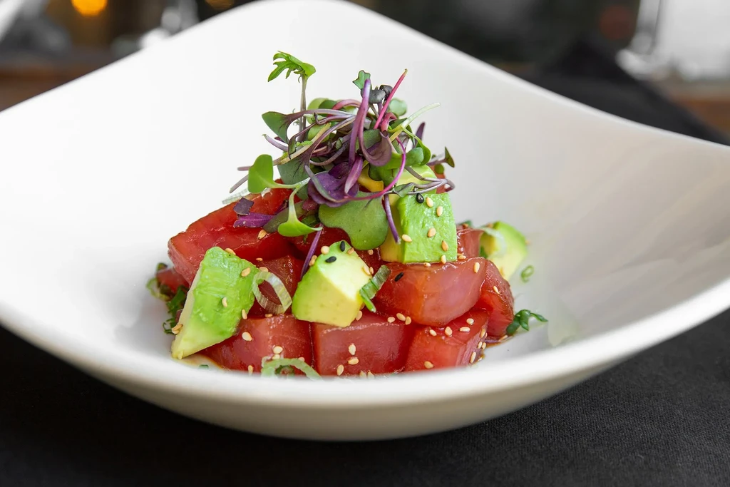 Cubed tuna and avocado poke with microgreens and sesame in white bowl.