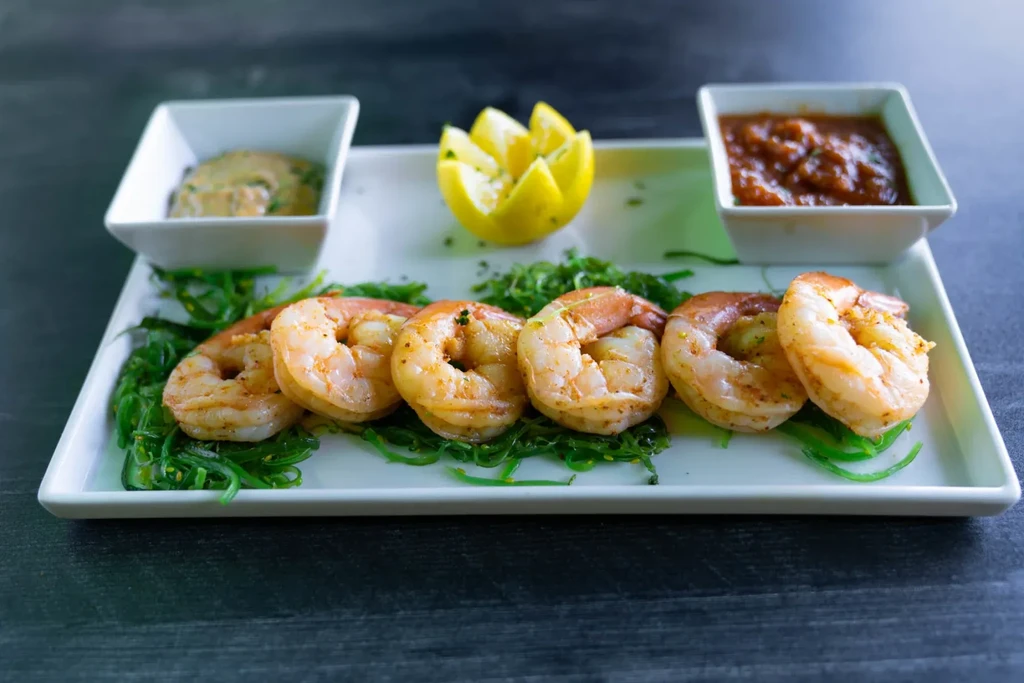 Cooked shrimp on green garnish, two sauces, and lemon on a white plate.