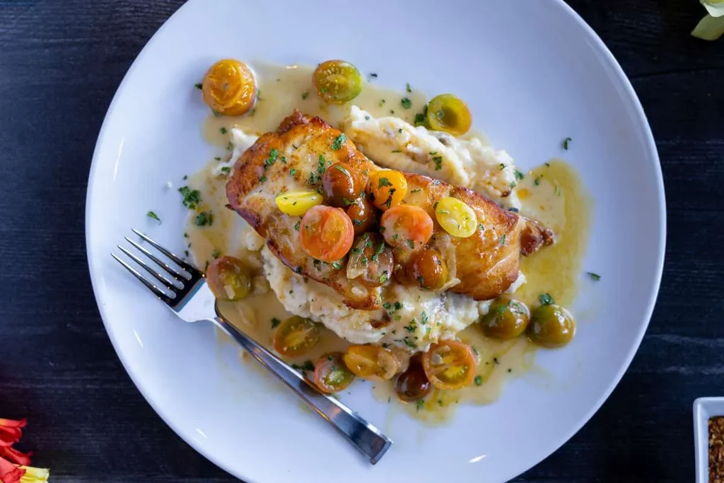 Golden-brown protein on purée, colorful tomatoes, herbs, and a fork.