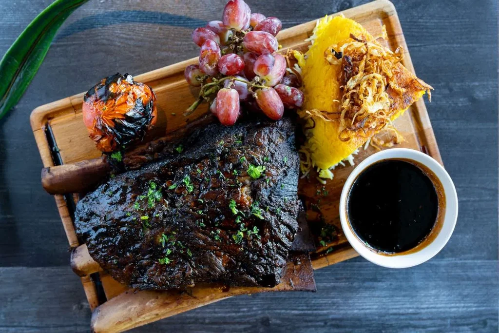 Top-down: Glazed meat, yellow rice, roasted tomato, grapes on a wood board.