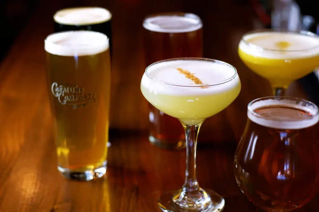 Six diverse cocktails and beers arranged on a polished wooden bar.