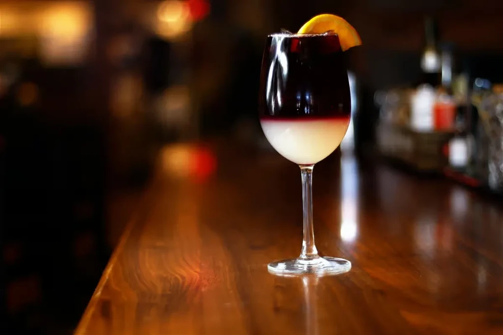Layered purple and white drink in stemmed glass, garnished with orange slice.