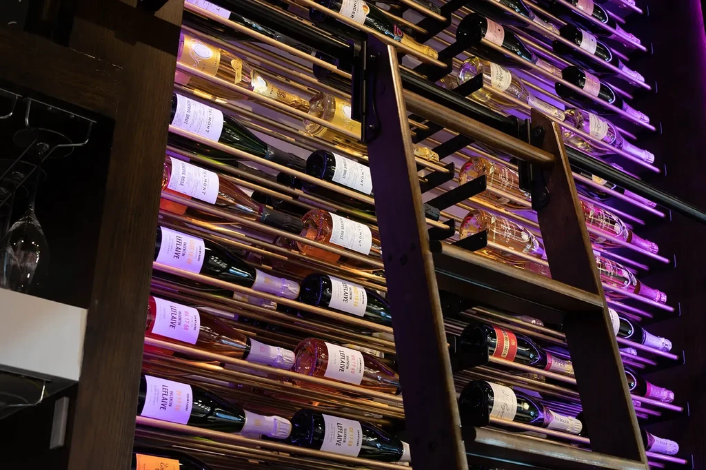 Purple-lit wine wall, bottles on metal racks, with a wooden ladder.