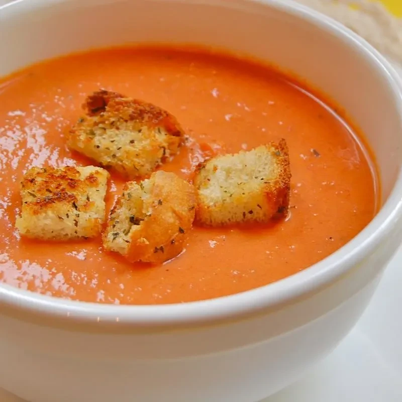 Orange soup with four herb-seasoned croutons in a white bowl.