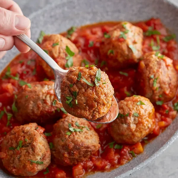 Hand lifting a herb-garnished meatball from chunky red tomato sauce.