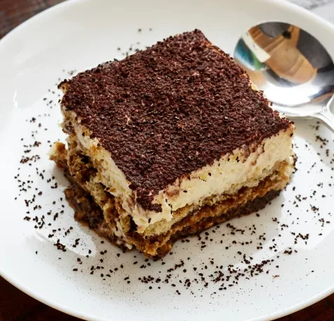 A slice of layered tiramisu on a white plate, dusted with cocoa.