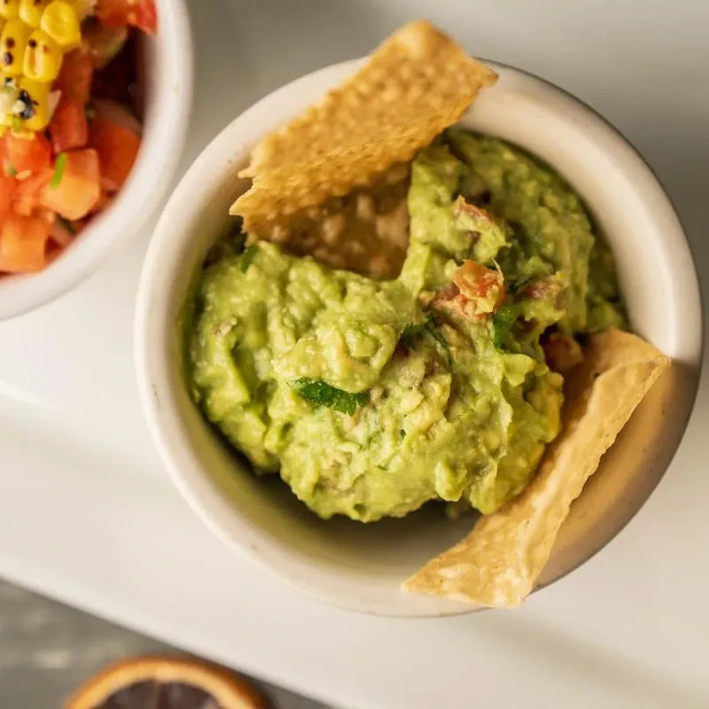 Chunky guacamole in a white bowl with tortilla chips and corn salsa.