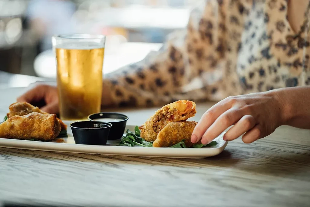 Plate of fried rolls, dipping sauces, and a beer, hand reaching.
