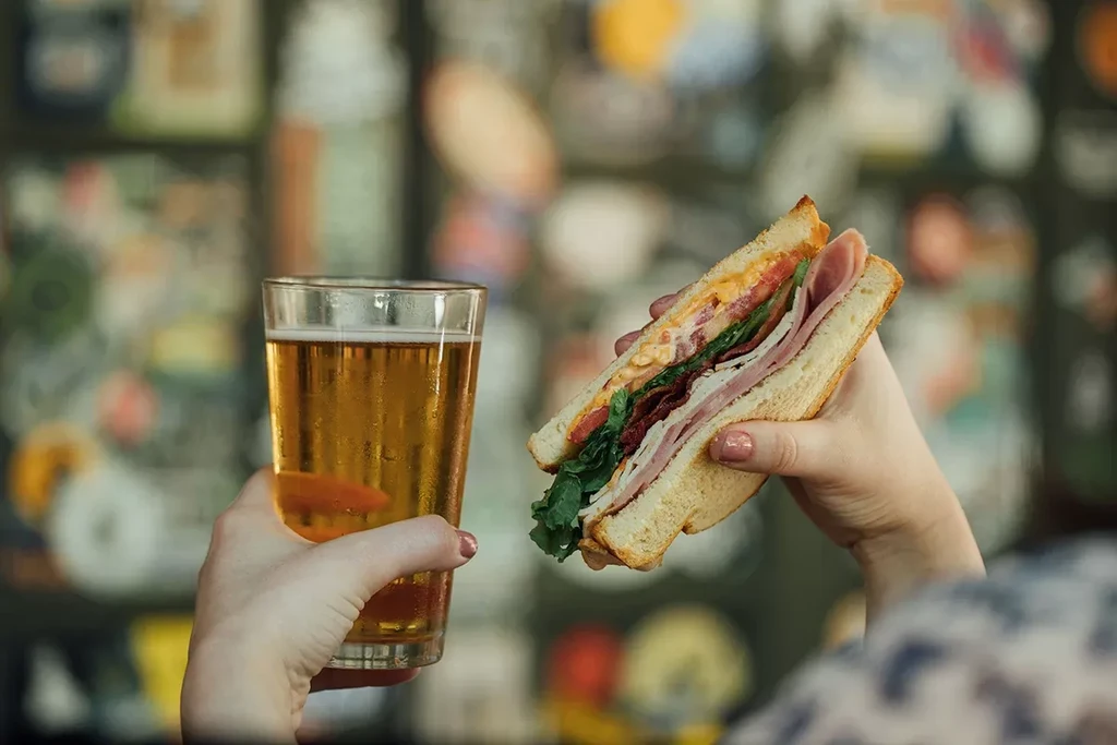 Hands holding a cold beer and a multi-layered club sandwich.