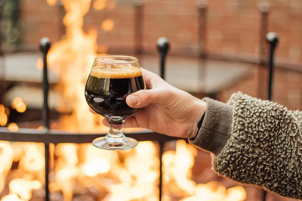 Hand holding dark beer snifter in front of a warm fire.