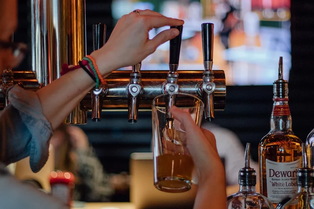 Close-up: hands pour beverage from tap into glass, blurred bar background.