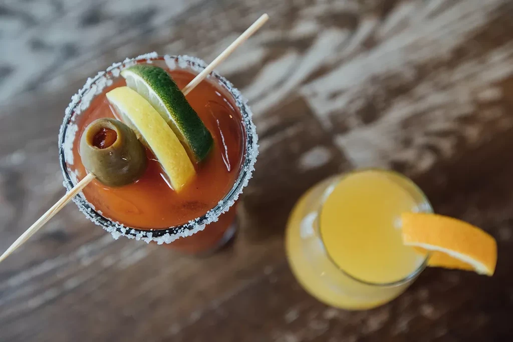 Top-down: Bloody Mary with garnishes and a yellow drink on wood.
