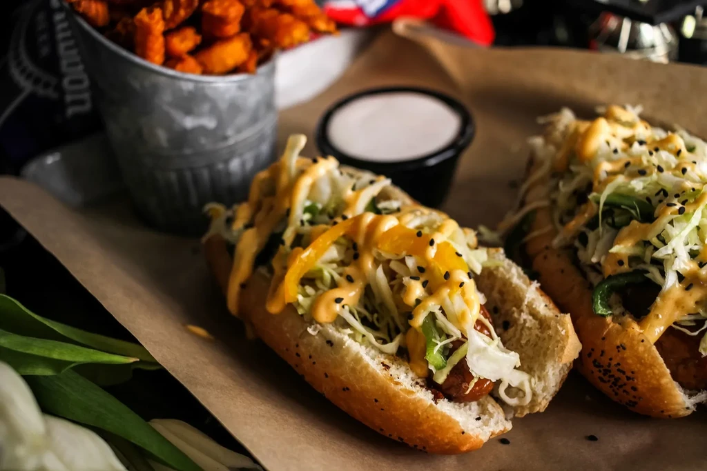 Two hot dogs topped with coleslaw, bell peppers, sauce, and sesame.