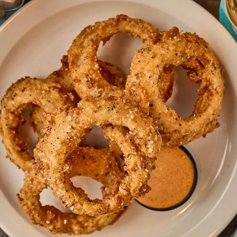 Seasoned golden-brown onion rings with orange dipping sauce on white plate.