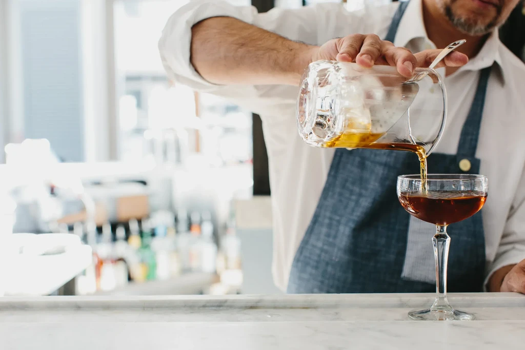Close-up of a bartender pouring a dark amber cocktail into a coupe glass.