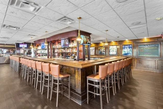 Empty U-shaped bar with stools, liquor, TVs, and framed sports jerseys.