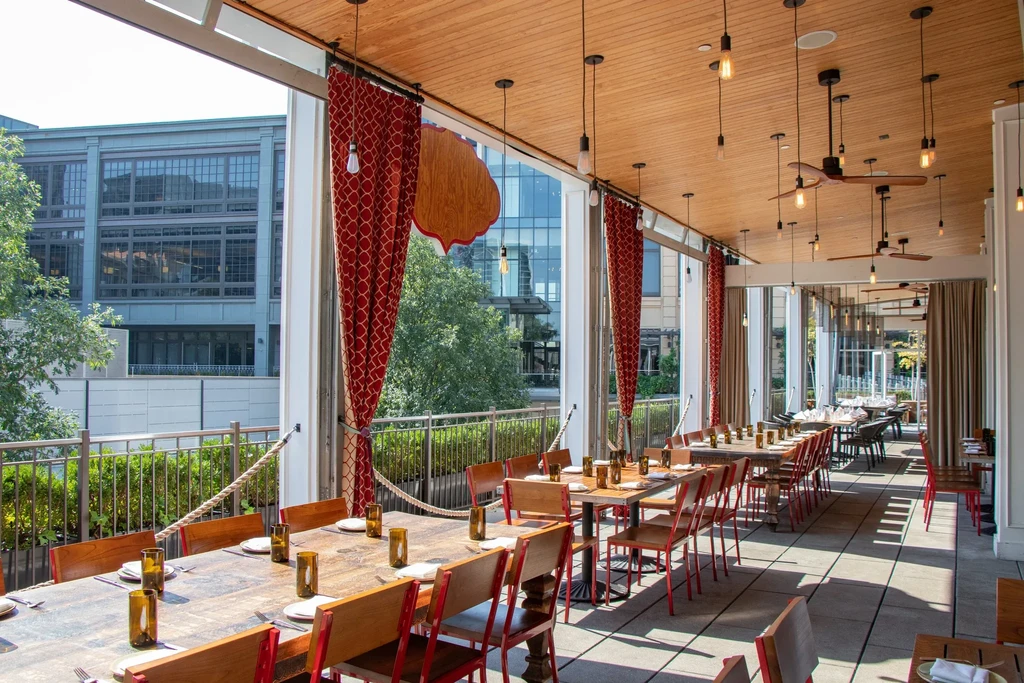 Modern restaurant patio with wooden tables, red-framed chairs, city view.