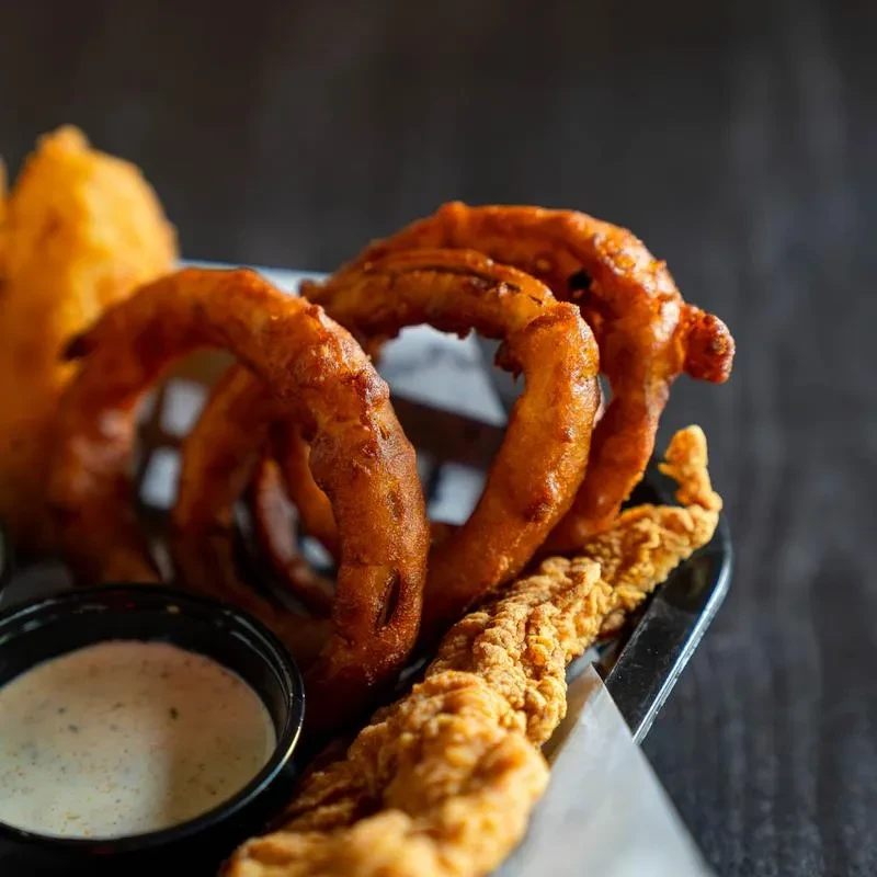 Golden-brown onion rings, fried chicken, and dipping sauce in a basket.